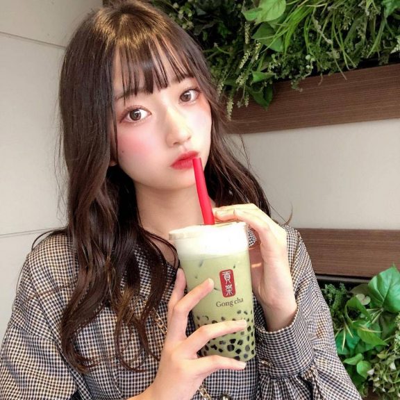 タピオカミルクティーの有名店「Gong Cha(ゴンチャ)」のタピオカ抹茶ミルクティーを飲みながらこちらをみている黒髪ロングヘアの女性。チェックのワンピースを着ている。